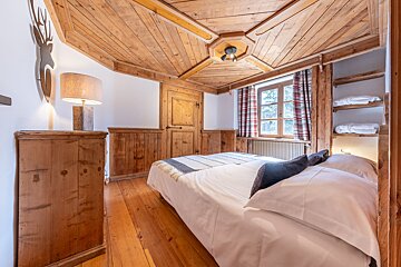 A cozy, rustic bedroom with extensive wood paneling, floor, and ceiling, featuring a comfortable bed, window with plaid curtains, and a deer silhouette decor.