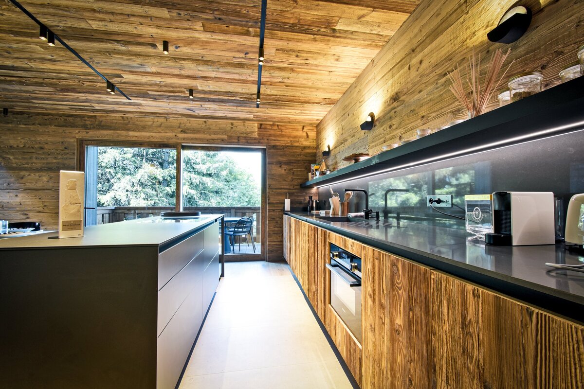 Sleek modern kitchen with rustic wooden walls/ceiling, dark island/counters, track lighting, and a large window overlooking a lush forest.