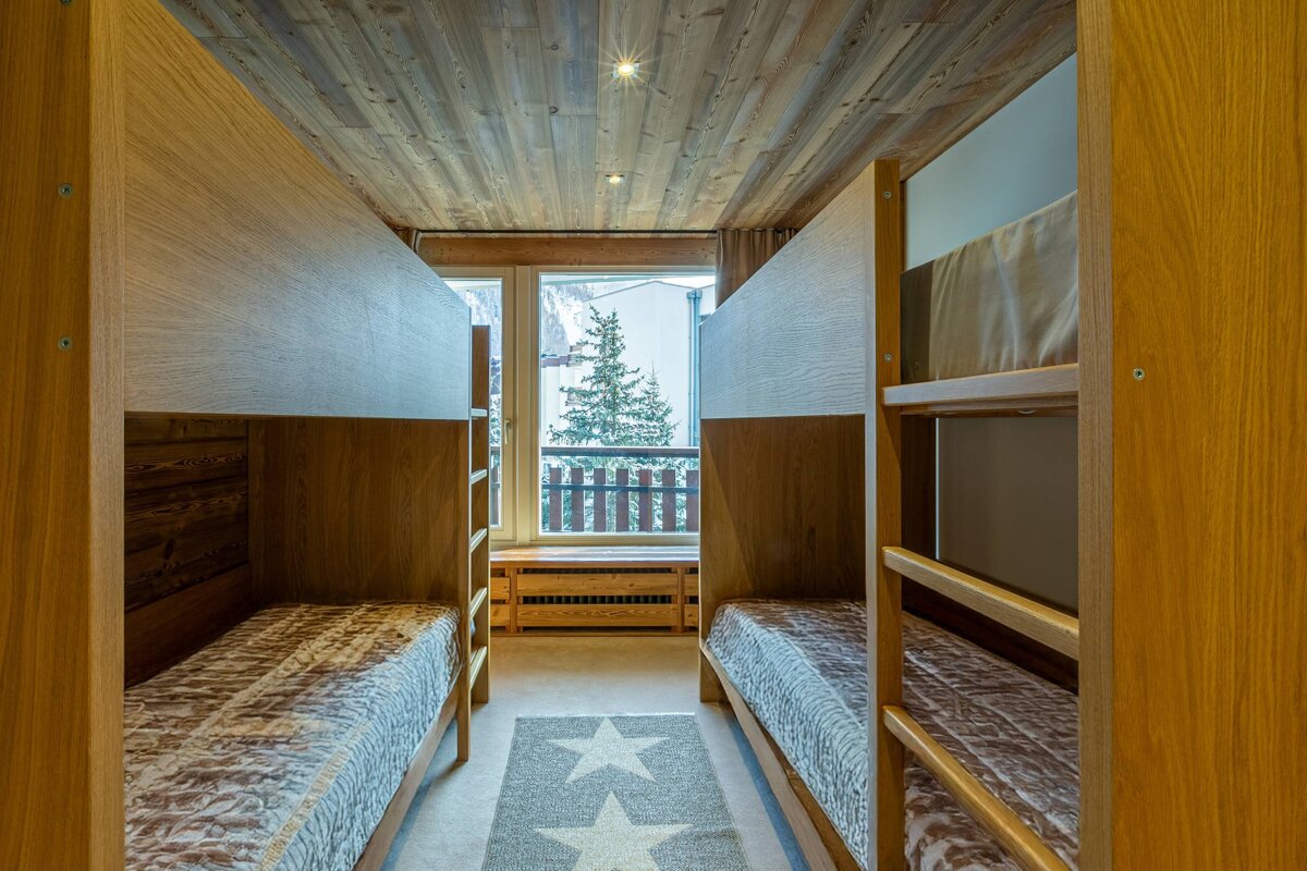 A rustic room with two wooden bunk beds, a wood-paneled ceiling, and a star rug. A window offers a snowy pine tree view.