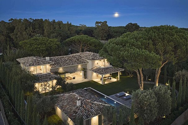 Aerial dusk shot of a sprawling luxurious villa with bright interiors, tiled roofs, a pool, and manicured gardens, nestled in a forest under a full moon.