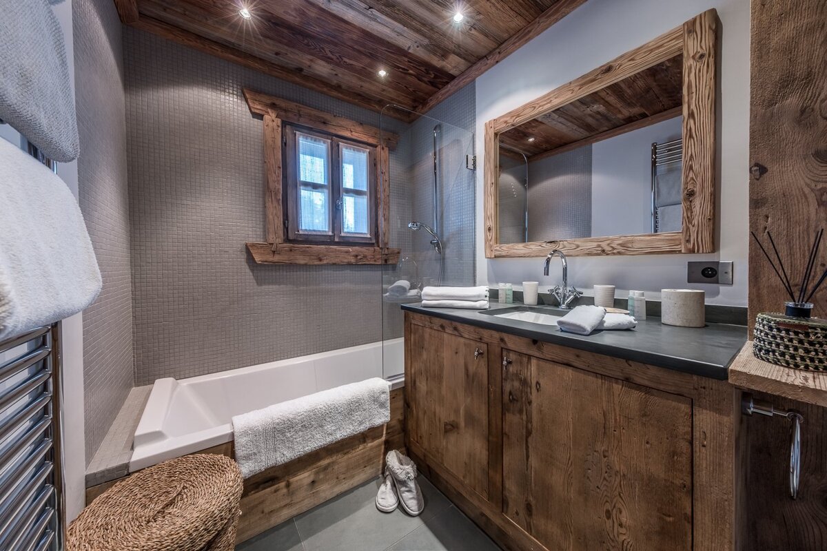 Rustic chalet bathroom featuring a wooden ceiling, vanity, mirror, and window frame. Includes a bathtub with shower and stylish grey tiled walls.