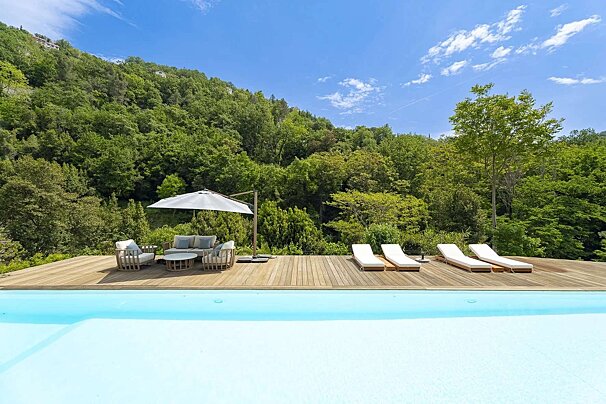 A serene infinity pool overlooks a wooden deck with modern outdoor lounge furniture and sunbeds, backed by a lush green forested hillside under a blue sky.