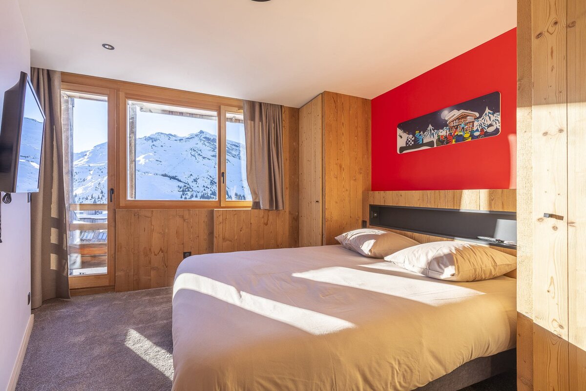 Cozy chalet bedroom featuring a large bed, red accent wall, wooden paneling, and a large window framing a scenic snowy mountain view.