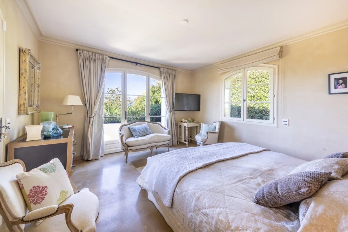 Elegant and bright bedroom with cream-colored walls, a large bed, armchairs, and French doors looking out to a garden.