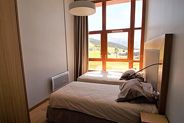 A bright bedroom with two single beds and a large window showcasing a stunning green mountain valley view.