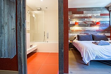 A rustic bedroom with a wood-plank accent wall next to an ensuite bathroom with bright orange floor tiles.
