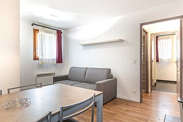 Simple living area featuring a grey sofa, a dining table in the foreground, white walls, wood floors, and an open doorway.