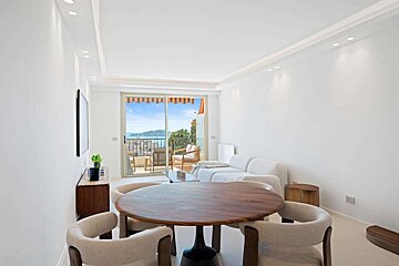 A bright, modern room features a round wooden dining table, white sofa, and a balcony with an orange awning overlooking the sea and a coastal town.