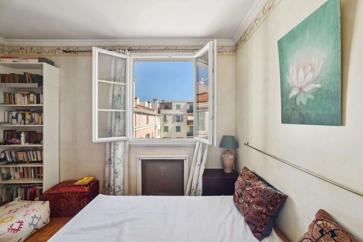 A cozy bedroom with an open window offering a view of city buildings. A bookshelf, bed with patterned pillows, and lotus artwork decorate the room.