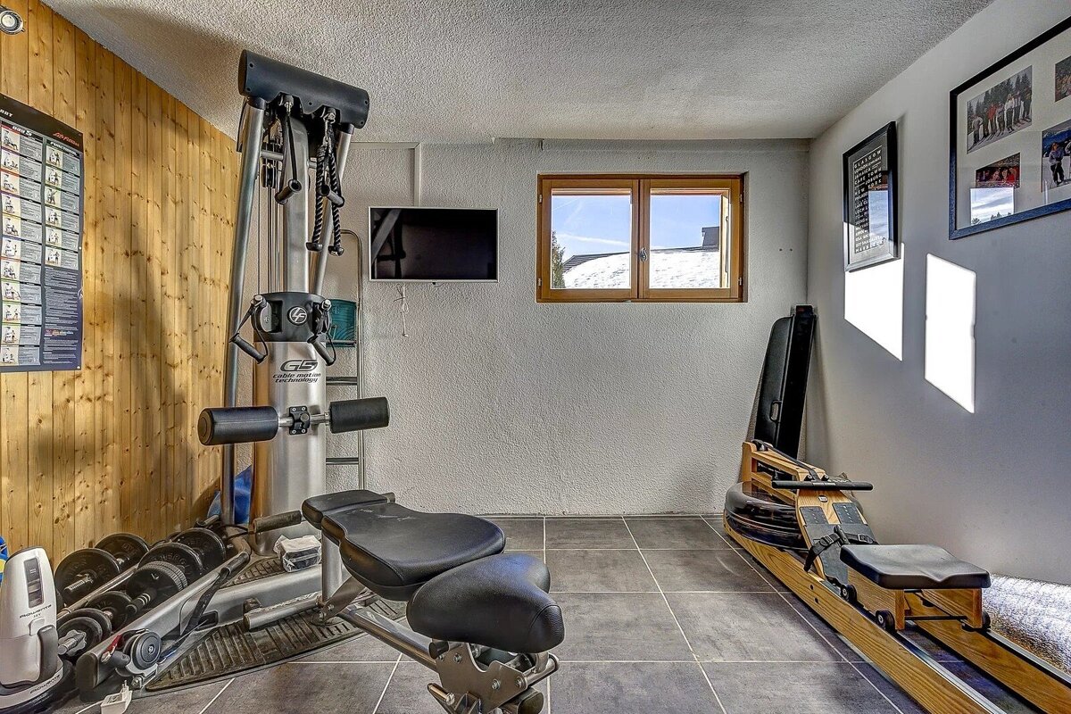 A compact home gym with a multi-gym, weights, rowing machine, and TV. A window offers a snowy view, complementing the wood-paneled and white walls.