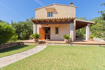 A charming tan two-story house with a covered patio, green lawn, and stone path under a clear blue sky, surrounded by trees.