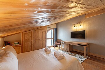 Cozy loft bedroom featuring a sloped wooden ceiling, a bed with white linens, built-in closets, and a desk with a wall-mounted TV.