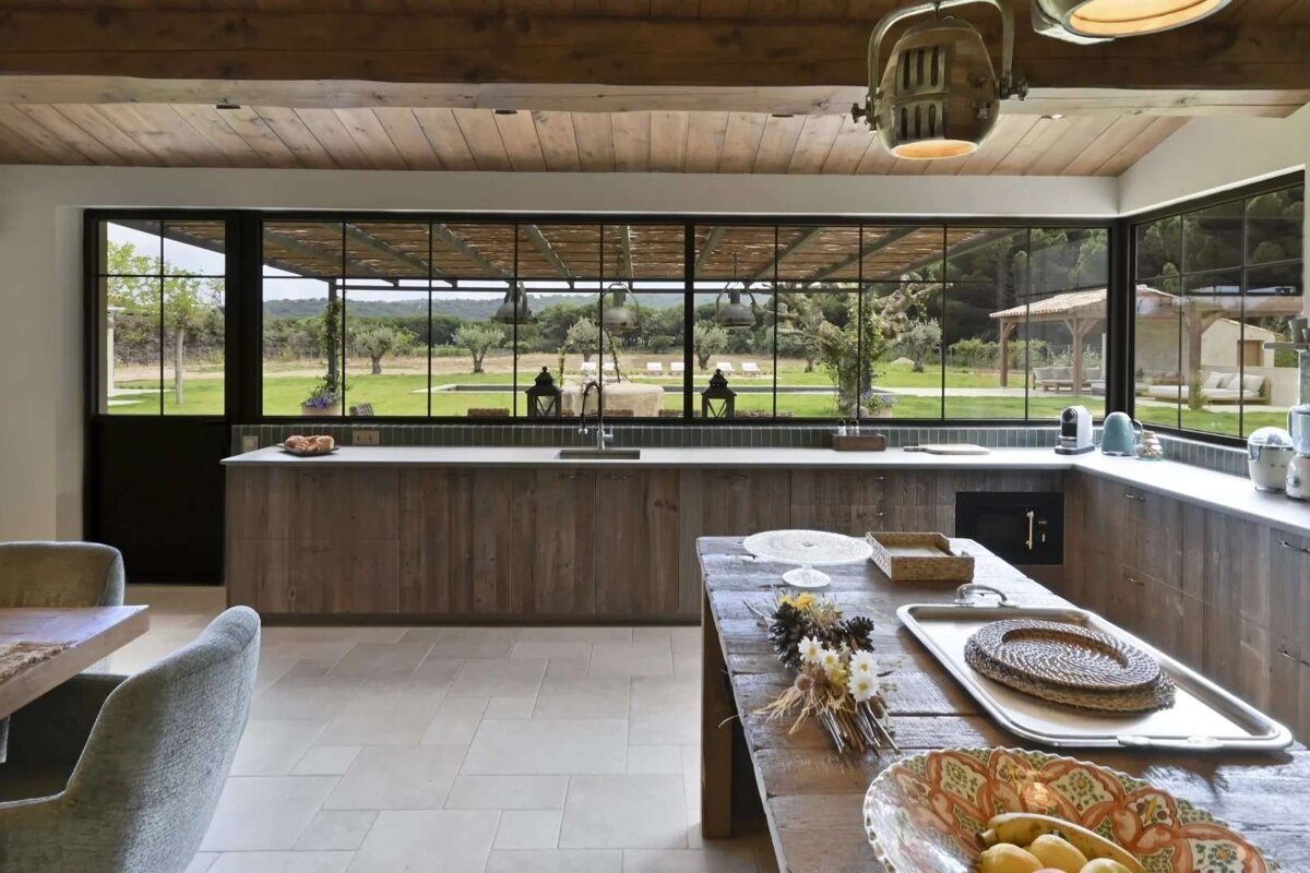 A rustic kitchen with wooden details and large window walls, providing a stunning view of a green landscape with olive trees, a pool, and outdoor seating.