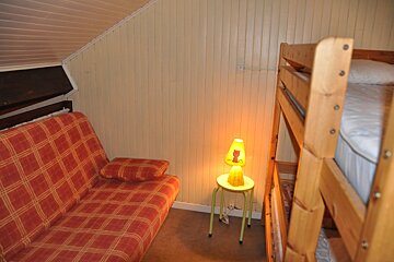 A cozy attic room featuring a red plaid futon, wooden bunk beds, and a yellow cat lamp on a stool. Paneled walls and a slanted ceiling.
