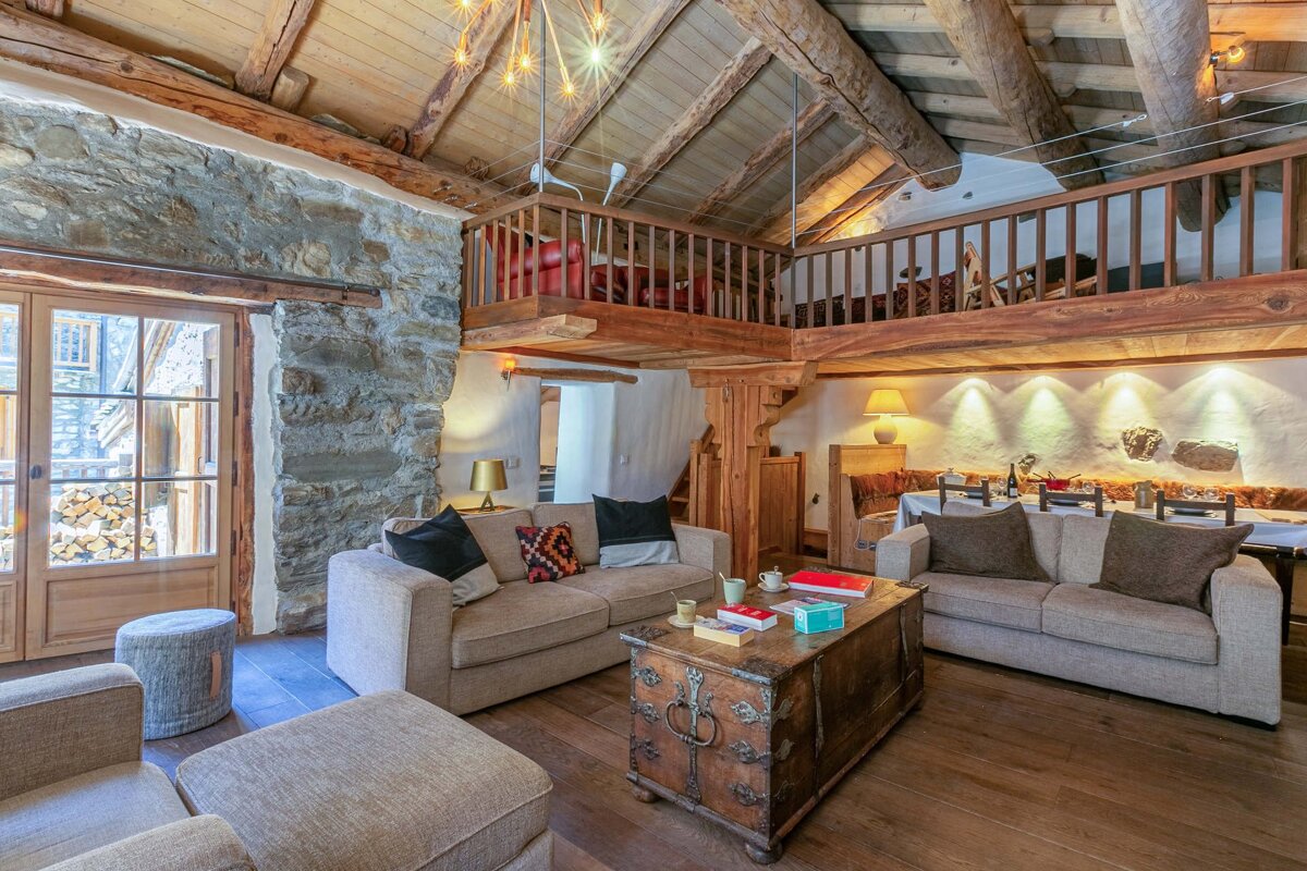 Cozy rustic cabin living room featuring stone walls, exposed wooden ceiling beams, a loft balcony, comfy sofas, and a vintage chest table.