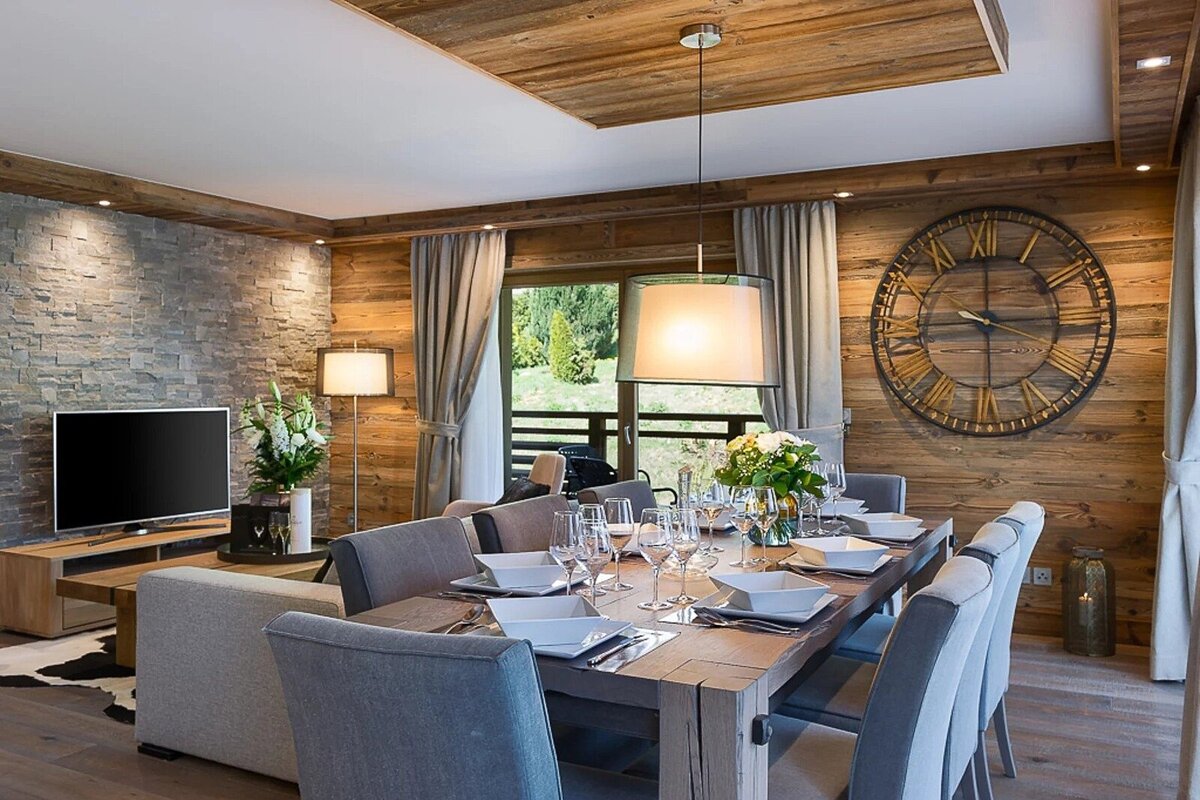 An elegant, rustic chalet living/dining room featuring a set wooden table, TV area with stone wall, wood paneling, and a large decorative clock.
