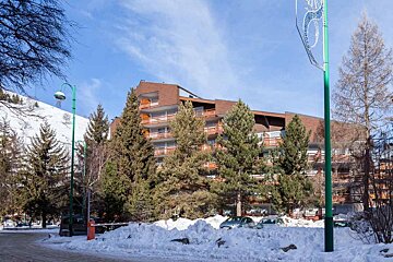 A large multi-story building, likely a hotel, is nestled among pine trees with snow on the ground, a snowy mountain, and a blue sky above.