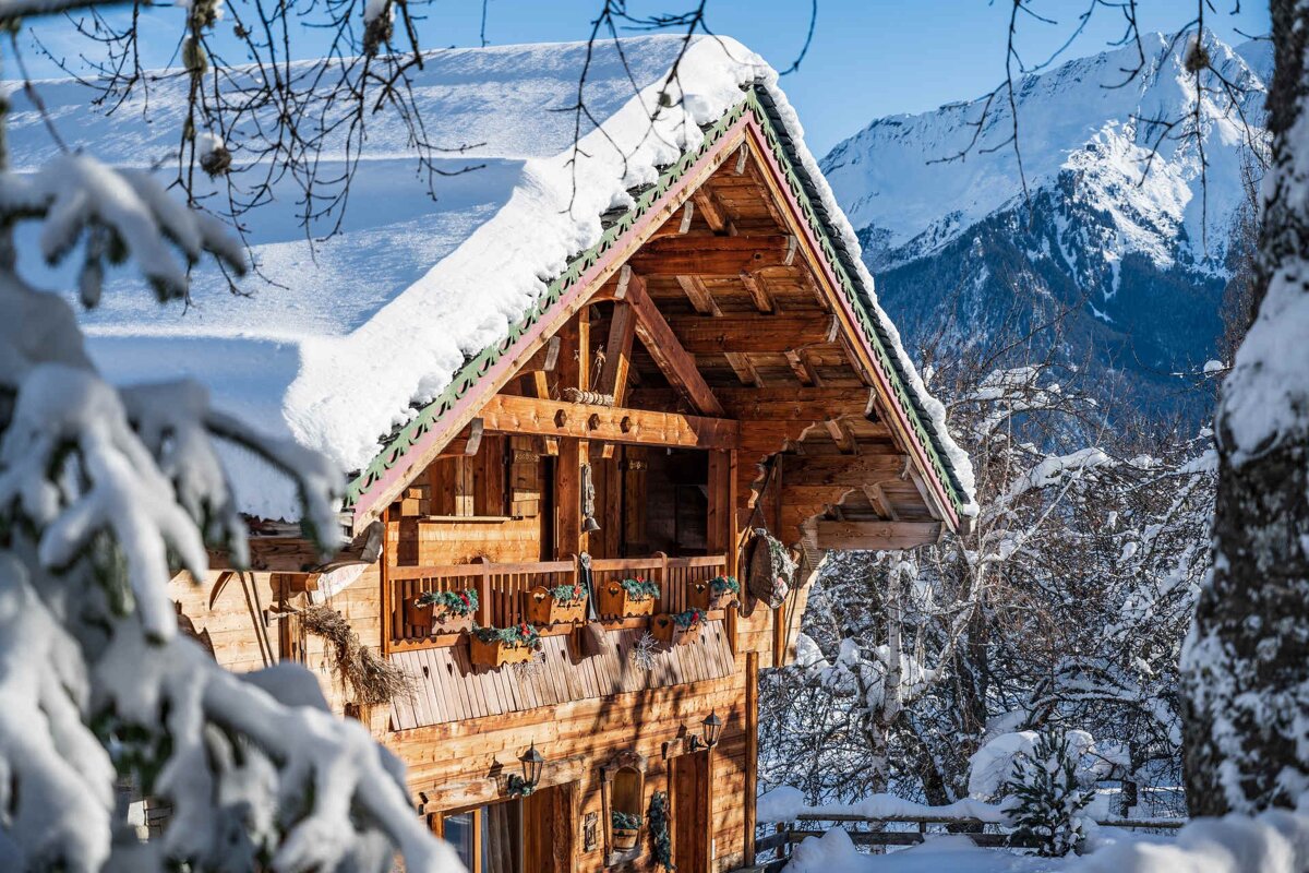 A beautiful wooden alpine chalet with a thick layer of snow on its roof, set against a backdrop of snow-capped mountains.
