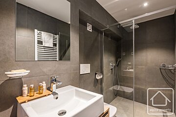 A modern bathroom with dark grey tiles, a white sink with toiletries, a large mirror, and a glass walk-in shower. A toilet is next to the sink.