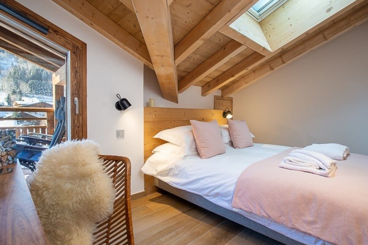 Cozy chalet bedroom under a slanted wooden ceiling with a skylight, featuring a bed with pink pillows and a balcony with a snowy view.