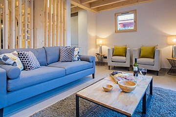 Cozy modern living room featuring a blue sofa, wooden coffee table with wine and snacks, and two armchairs with yellow pillows.