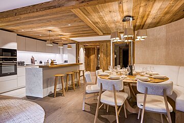 A modern open-plan kitchen and dining area with a rustic wooden ceiling. A large dining table is set for guests, beside a kitchen island with stools.