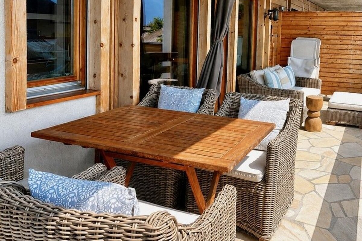 A cozy outdoor patio featuring a wooden dining table and wicker armchairs with blue patterned pillows on a stone-tiled floor.