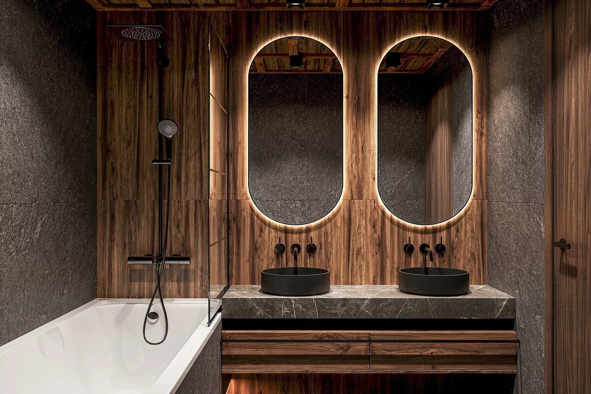 A modern bathroom with dark wood and gray stone walls, featuring a bathtub with shower, double vanity with black sinks, and illuminated oval mirrors.