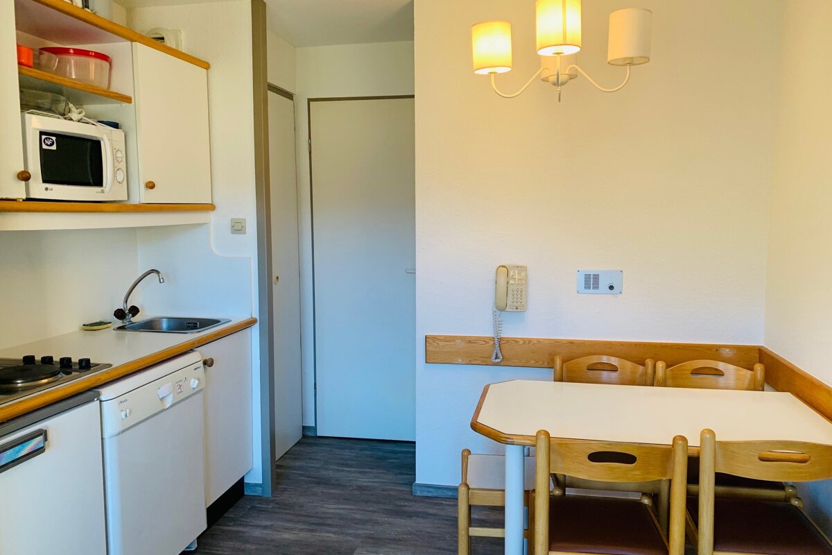 A compact kitchen and dining area in a studio apartment, featuring white cabinets, a microwave, sink, stove, and a small dining table with wooden chairs.