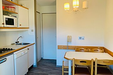 A compact kitchen and dining area in a studio apartment, featuring white cabinets, a microwave, sink, stove, and a small dining table with wooden chairs.