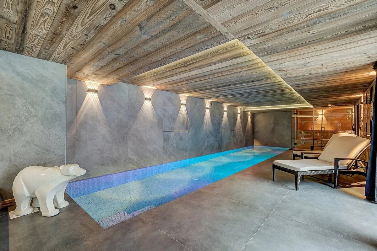 Modern indoor pool room with a long blue pool, textured wooden ceiling, grey walls, and elegant lighting. A white polar bear statue stands by the pool.
