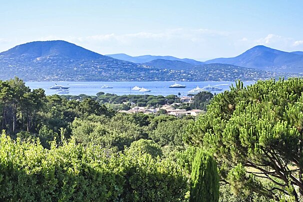 Scenic view of a Mediterranean bay with yachts, framed by green pine trees and rolling blue hills in the distance.