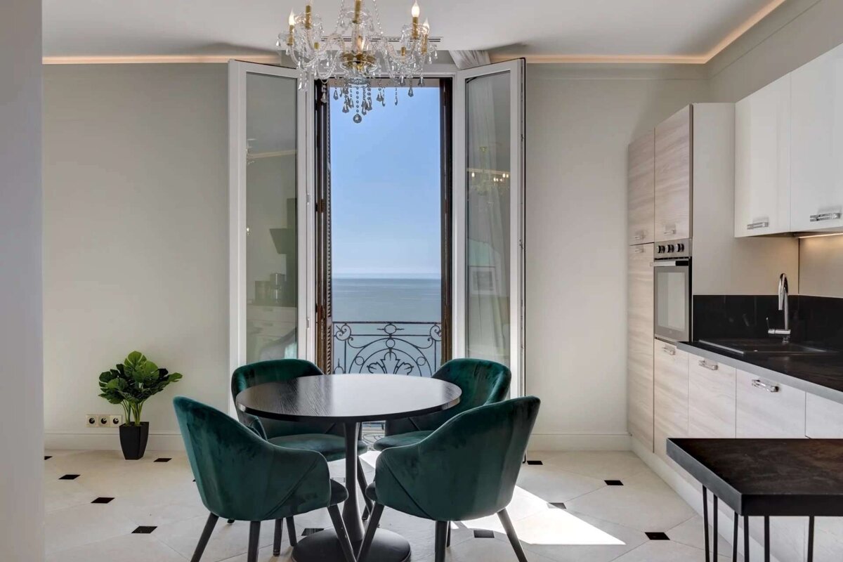 A modern kitchenette and dining area with green velvet chairs and a black table, illuminated by a chandelier, opens to a balcony overlooking the sea.