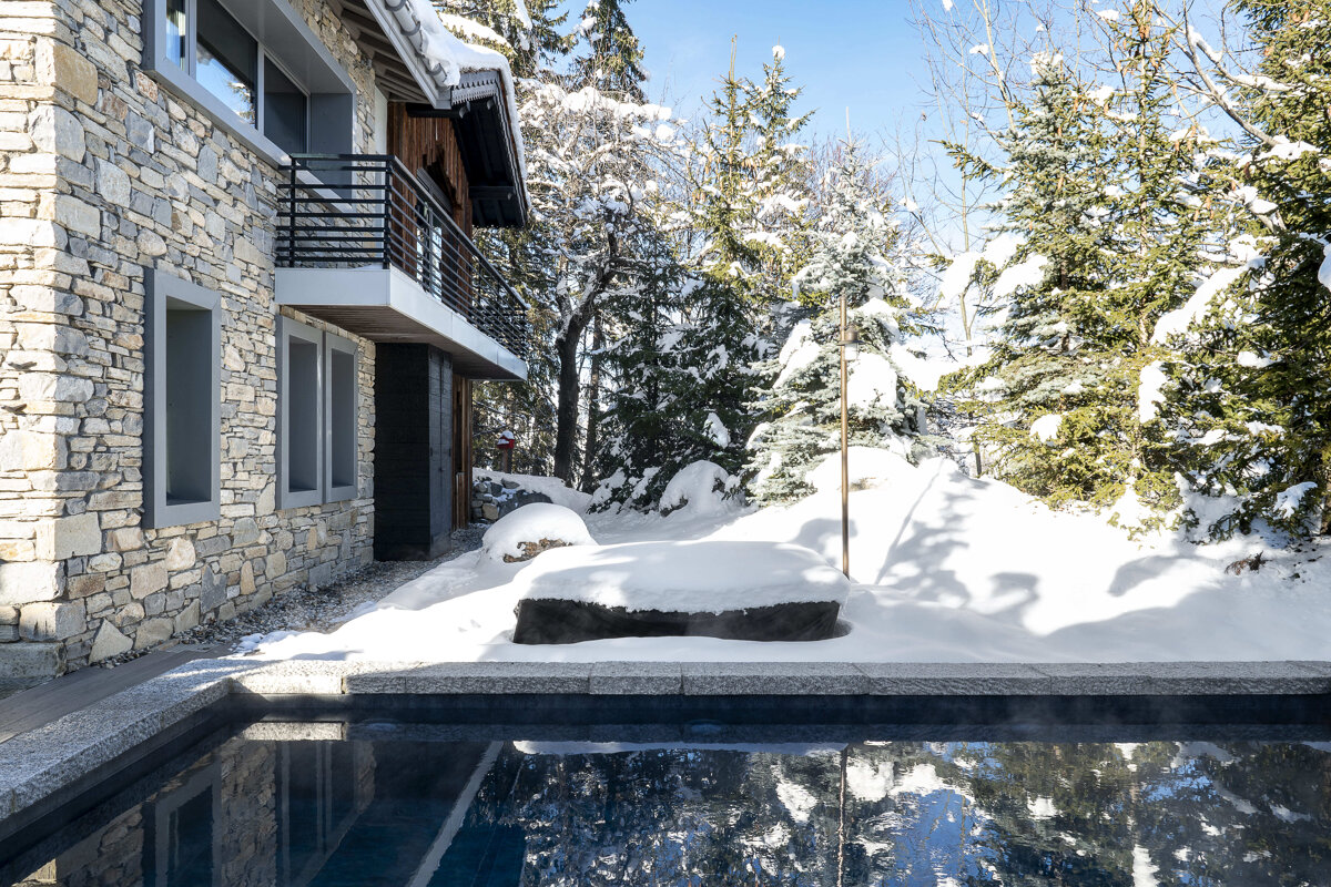 A stone house with a balcony next to a steaming outdoor pool, surrounded by snow-covered pine trees in a picturesque winter landscape.