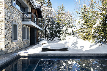 A stone house with a balcony next to a steaming outdoor pool, surrounded by snow-covered pine trees in a picturesque winter landscape.