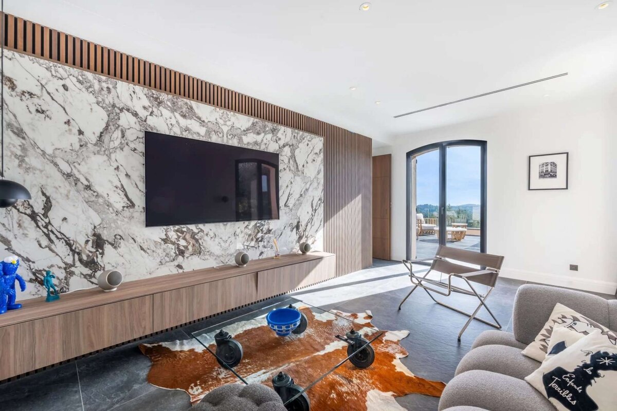 A modern, bright living room features a marble accent wall with a TV, wood slatted panels, and a glass coffee table over a cowhide rug.
