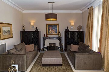 An elegant living room features two brown sofas, a central ottoman, and a fireplace flanked by dark cabinets. Beige walls, gold curtains, and classic art complete the look.