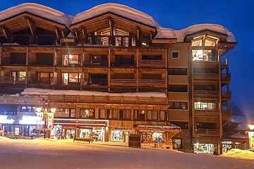 A large, snow-covered wooden chalet building at night with illuminated windows, shops, and restaurants on the ground floor. Snow covers the roofs and ground.