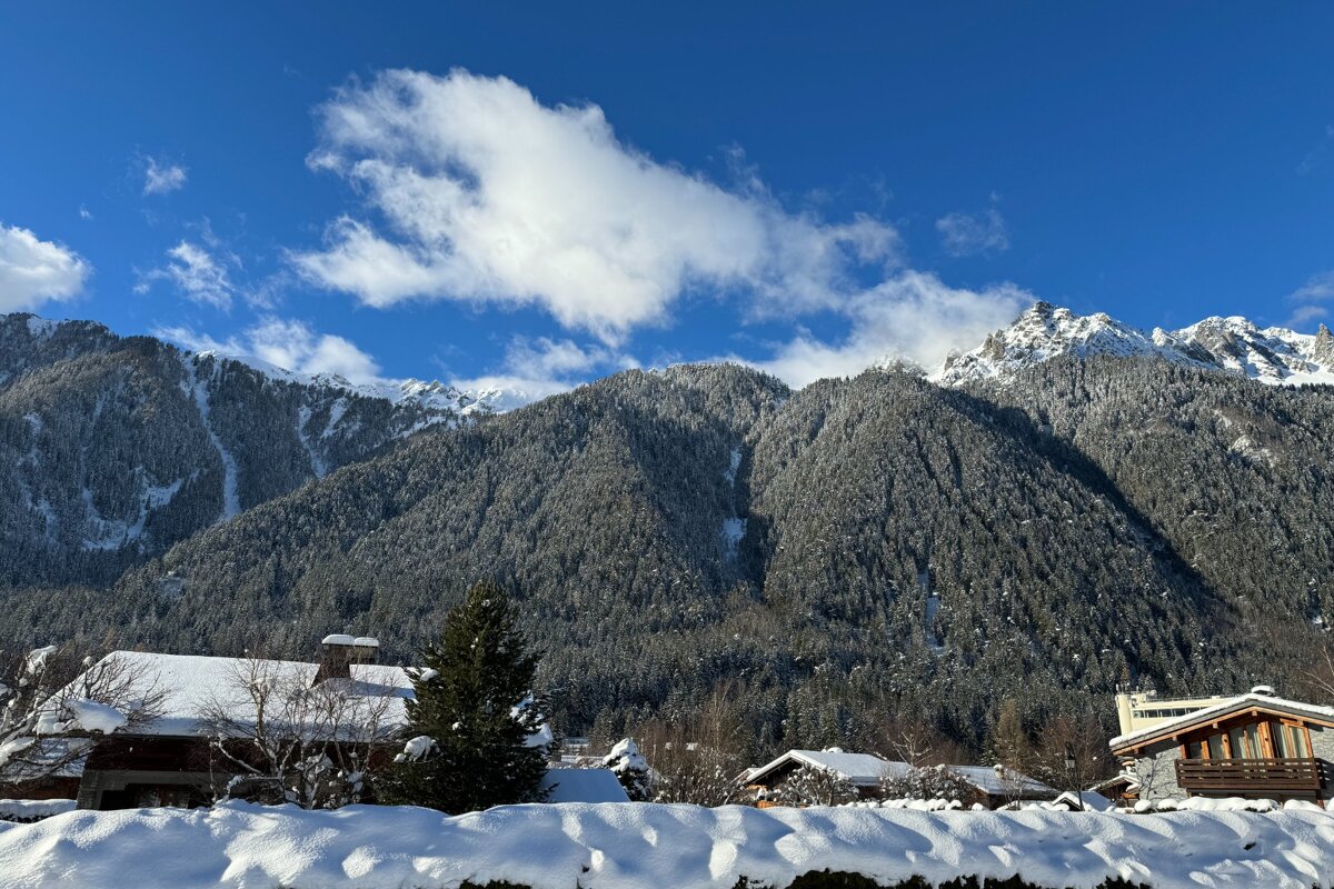Snow-covered mountains with dense evergreen trees, houses, and a bright blue sky with white clouds create a serene winter landscape.