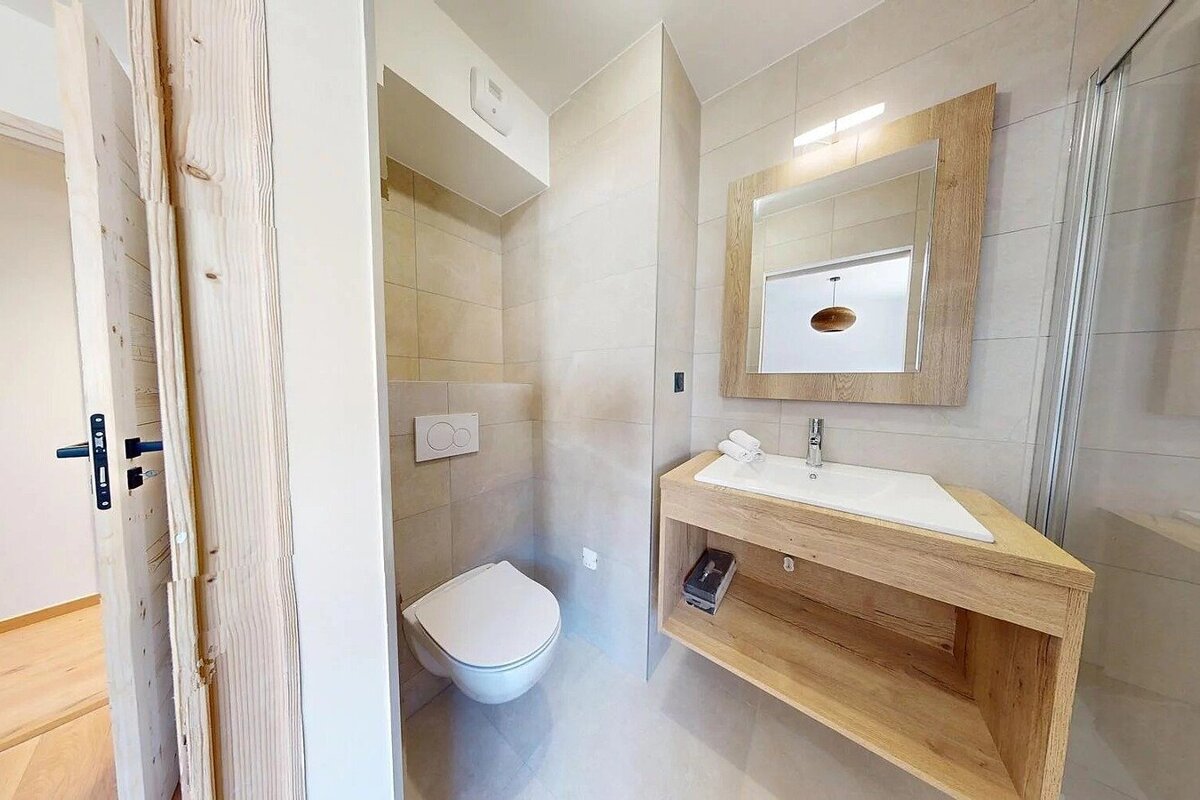 A modern bathroom features light-colored tiles, a wall-mounted toilet, and a wooden vanity with a sink and mirror. A shower is partially visible.