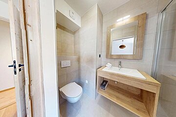 A modern bathroom features light-colored tiles, a wall-mounted toilet, and a wooden vanity with a sink and mirror. A shower is partially visible.