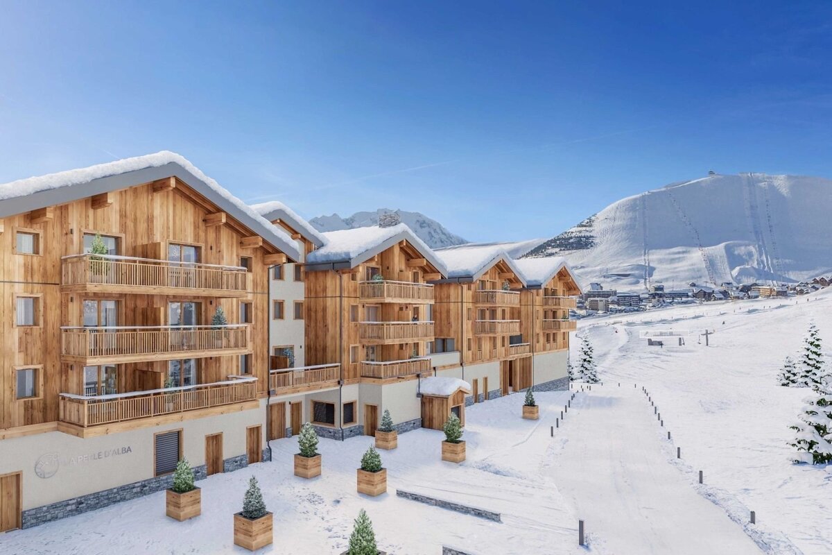 Snow-covered wooden chalets at a ski resort, set against a backdrop of snowy mountains under a clear blue sky.