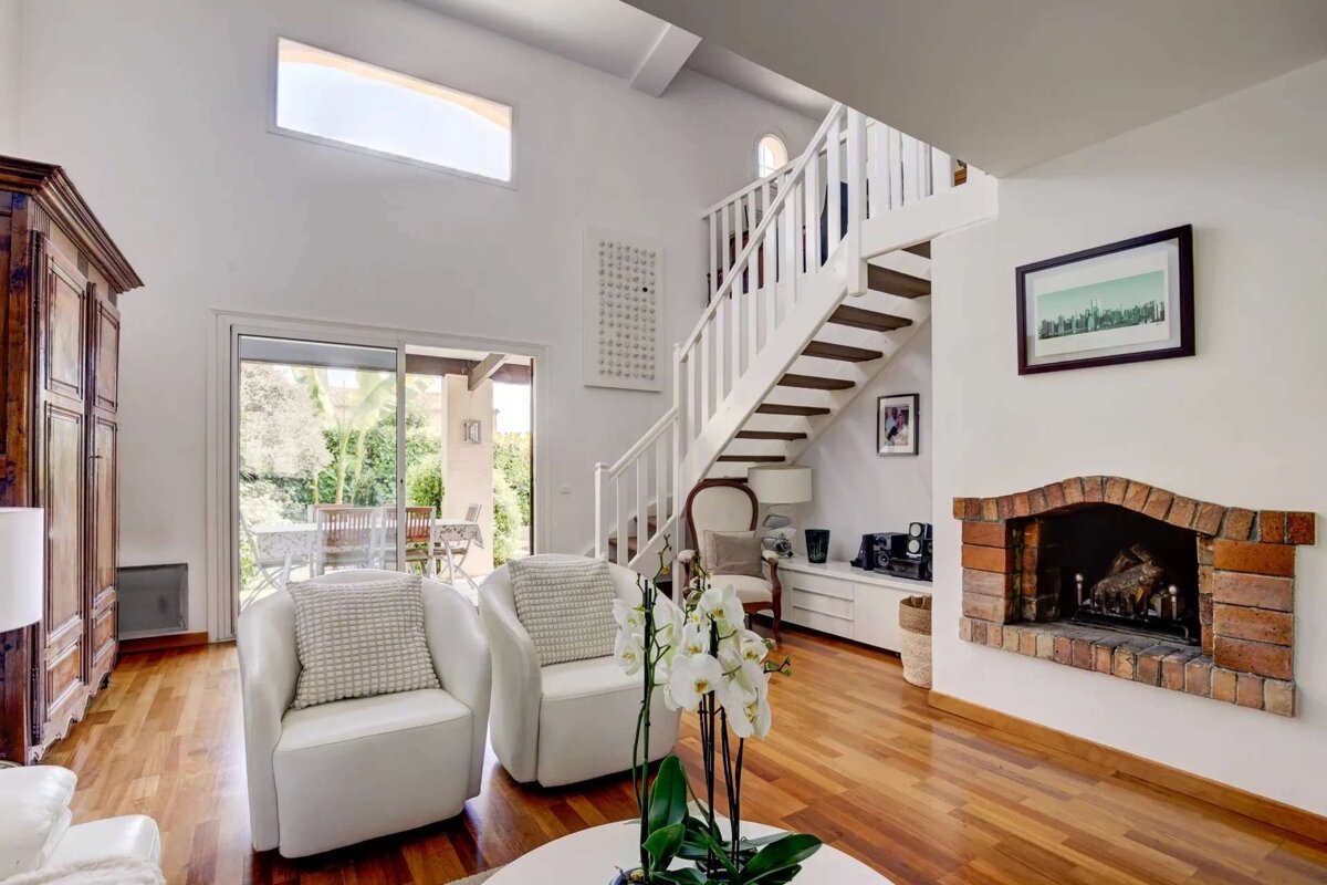 Bright living room with white armchairs, wooden floor, a white staircase, and a brick fireplace. Sliding doors lead to an outdoor patio.