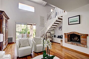 Bright living room with white armchairs, wooden floor, a white staircase, and a brick fireplace. Sliding doors lead to an outdoor patio.