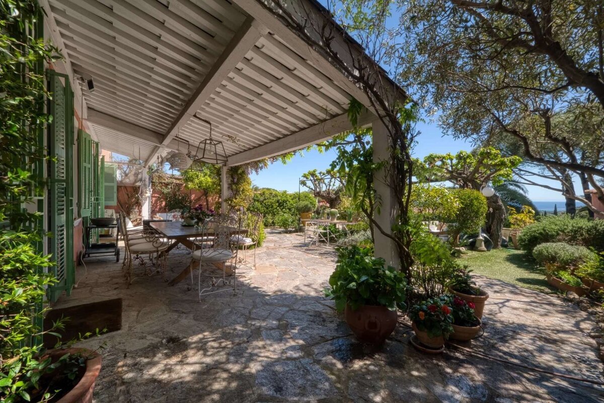 A beautiful covered outdoor patio with a dining area and stone floor, surrounded by lush green gardens, leading to a distant ocean view under a clear sky.