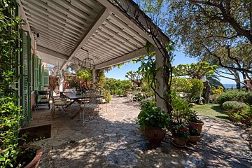 A beautiful covered outdoor patio with a dining area and stone floor, surrounded by lush green gardens, leading to a distant ocean view under a clear sky.