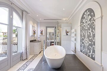 Elegant, light-filled bathroom featuring a freestanding tub, ornate mosaic wall, French doors to a balcony, and modern art.