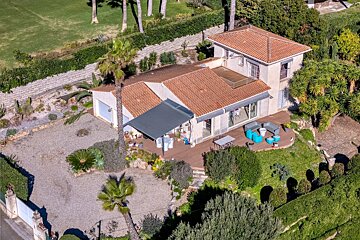 High-angle shot of a sunny Mediterranean house featuring a tiled roof, outdoor patio with blue furniture, and surrounding greenery.