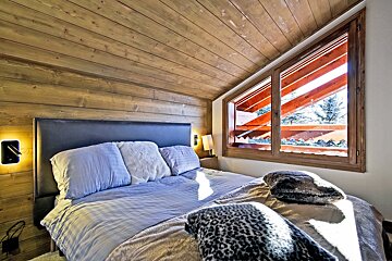 Cozy alpine bedroom with wood-paneled walls, a slanted ceiling, a plush bed, and a large window looking out at snowy trees.