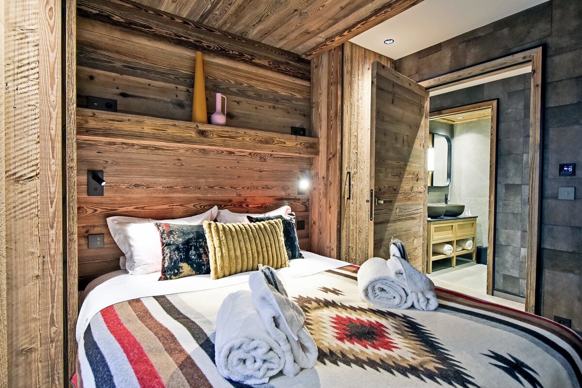 Cozy, rustic chalet bedroom with wood-paneled walls and an open door leading into a modern stone bathroom.